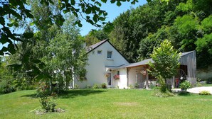 Ein Haus mit weißer Fassade, einer Veranda mit Sitzgelegenheiten und einem an ein Holz-Stadel erinnernden Bau auf der rechten Seite.