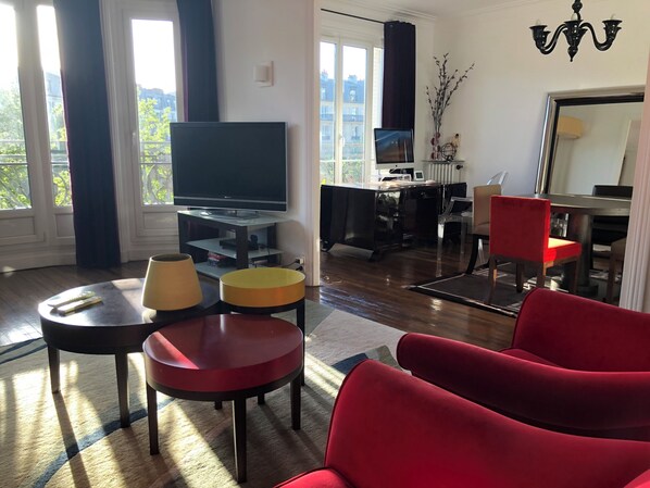 Una sala moderna con televisor de pantalla plana, un comedor con mesa y sillas, y vistas a la ciudad a través de amplias ventanas.