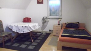 Quarto pequeno e simples com uma cama, uma mesa, uma cadeira, um radiador e uma janela.