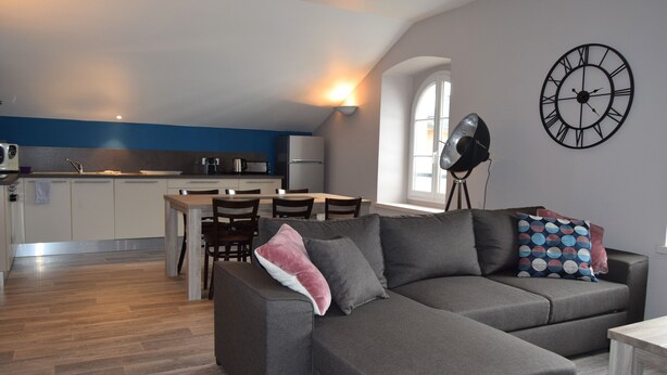 Une cuisine moderne avec des meubles blancs, un coin repas comprenant une table et des chaises, et un canapé gris agrémenté d’un coussin rose.