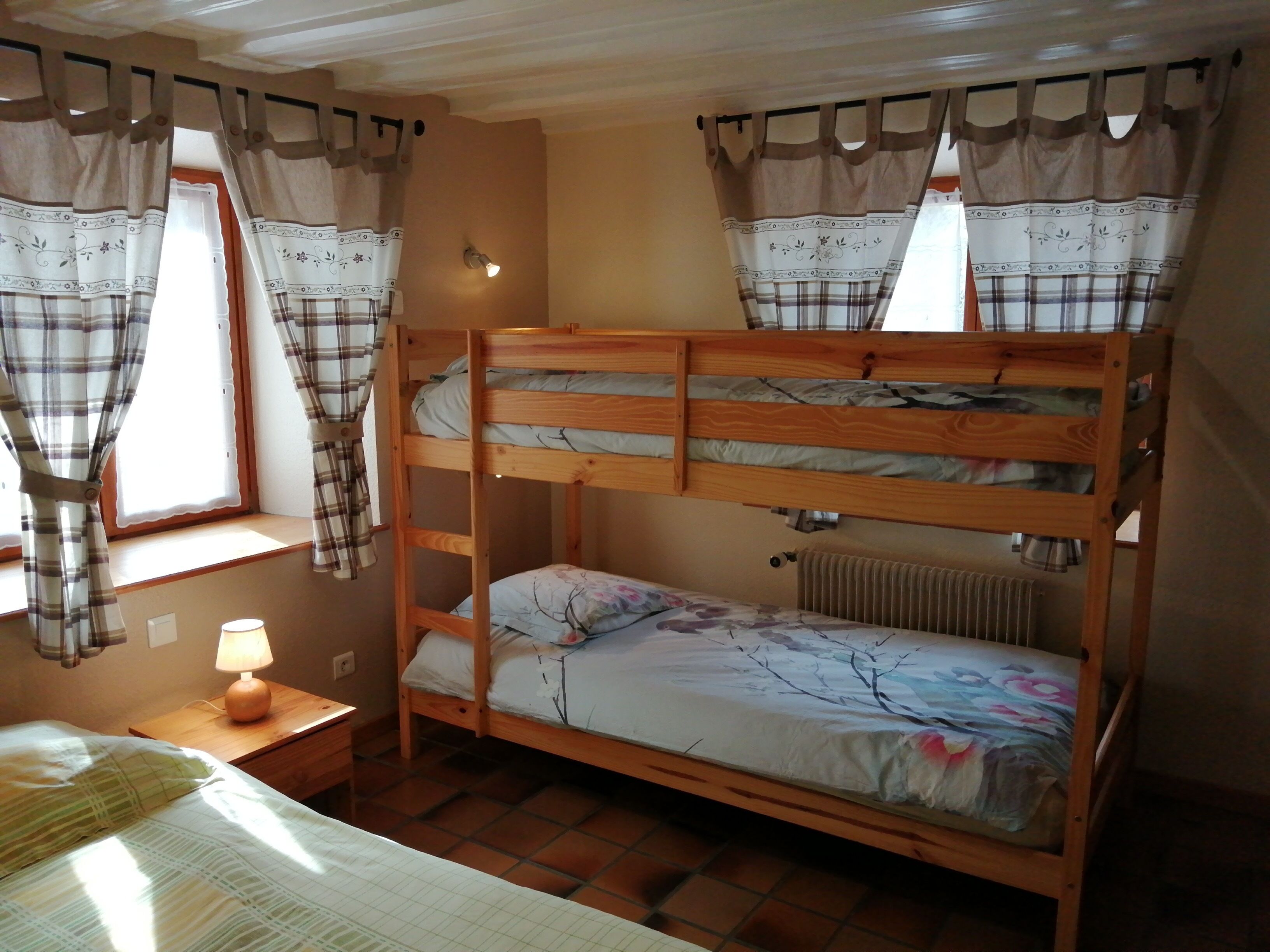 Una habitación con literas, una mesita de noche con lámpara, una ventana con cortinas y un radiador.
