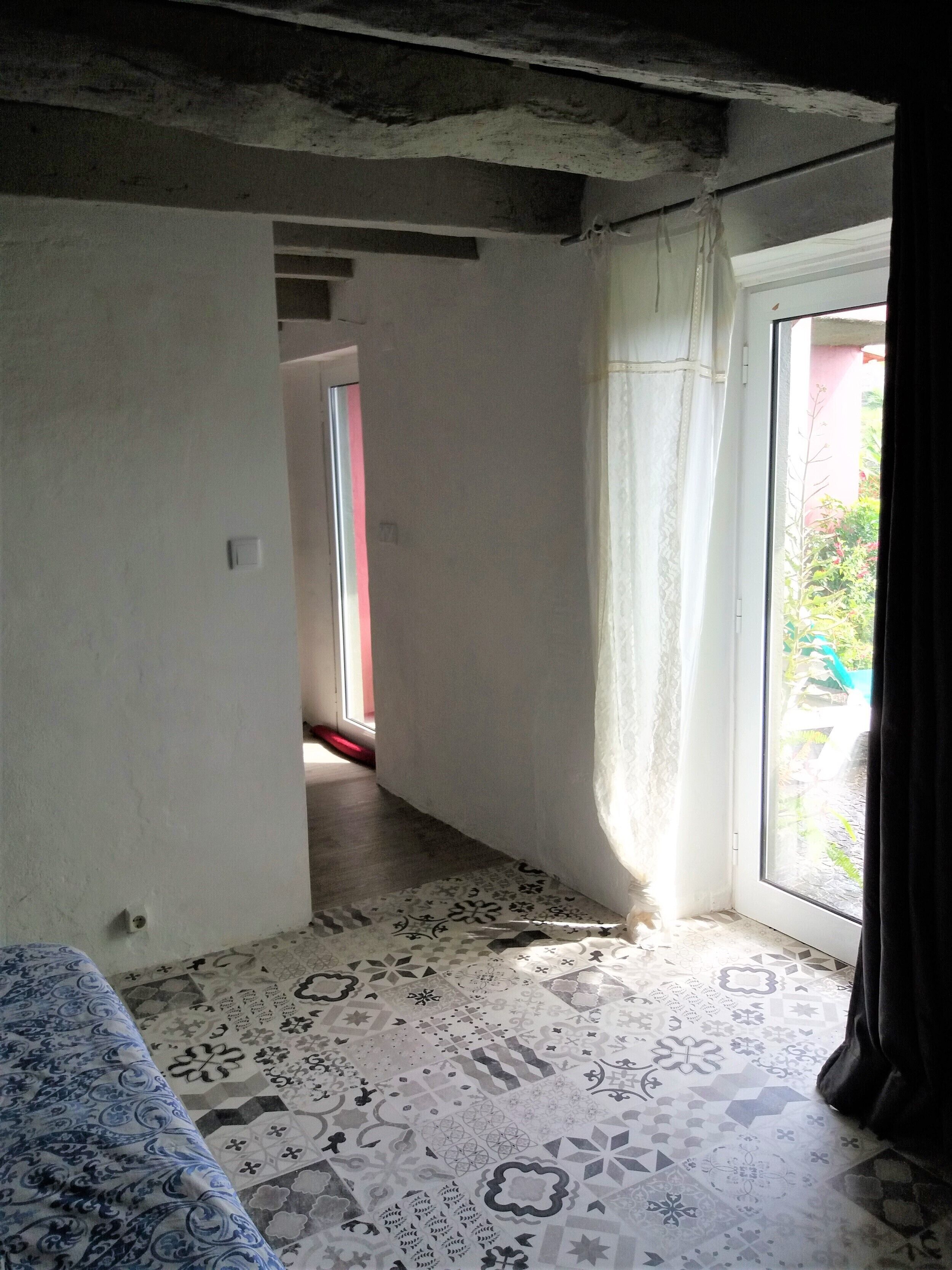 Une pièce avec un sol à motifs, un lit, une porte donnant sur une autre pièce et une vue par une fenêtre avec des rideaux.