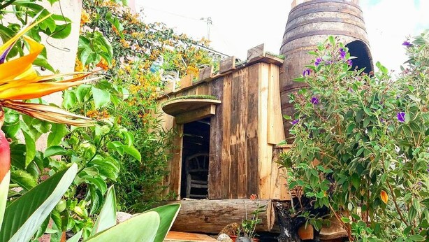 Una construcción rústica de madera con una pequeña entrada, rodeada de abundante follaje y un tanque cilíndrico grande al fondo.