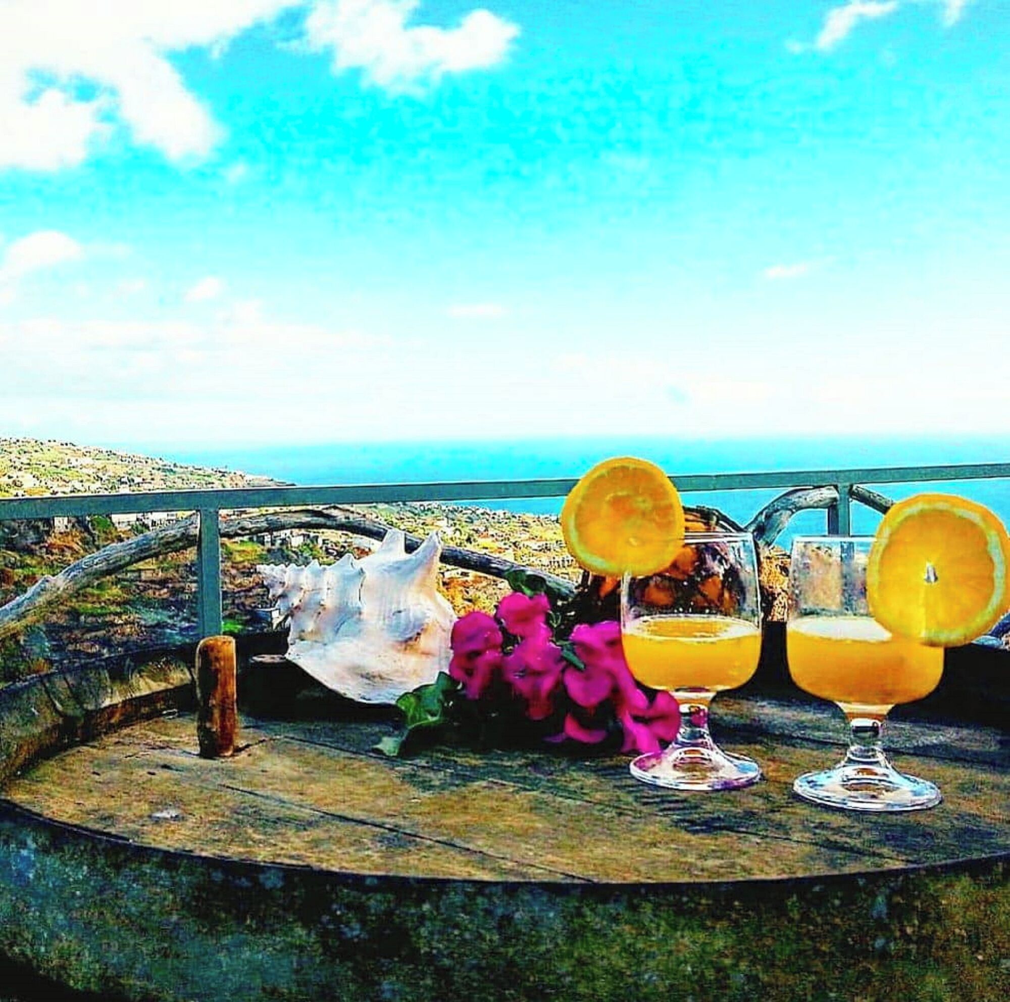 Ein malerischer Ausblick mit einem Holztisch, auf dem zwei Orangensäfte, eine große Muschel und rosa Blumen stehen.