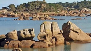 Un paisaje costero con grandes rocas, agua tranquila y una colina distante con edificios.