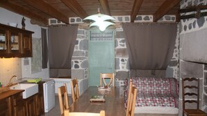 Una cocina rústica con una mesa de madera, sillas y un comedor. Se aprecia una pared de piedra, una puerta verde y una cama con colcha estampada.