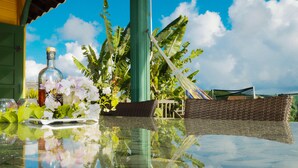 Una mesa de vidrio con una botella y flores, una cesta tejida y vistas de un edificio verde y plantas de banano.