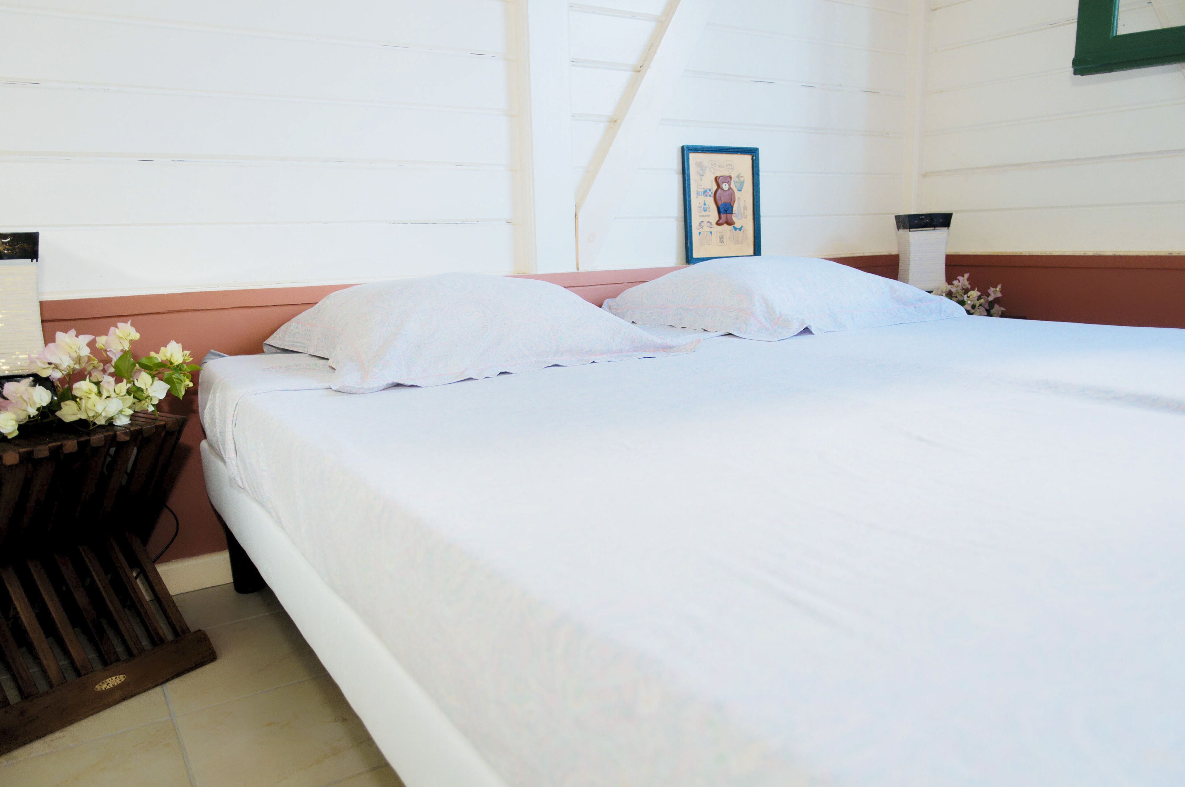A bedroom with two beds, a wooden nightstand with a vase of flowers, and a framed picture on the wall.