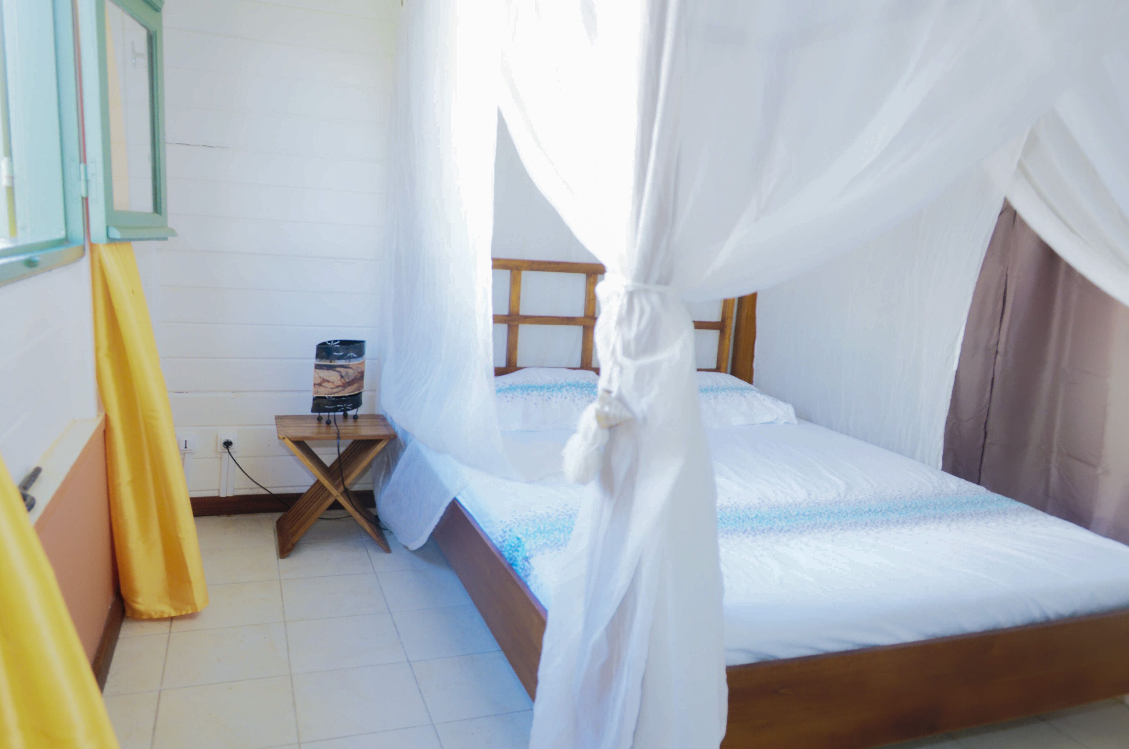 Una cama individual con estructura de madera y ropa de cama blanca, una mesita de noche con lámpara y una ventana con cortinas.