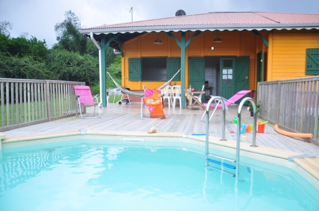 A pool area with a wooden deck, a covered seating area, and a building with green shutters.