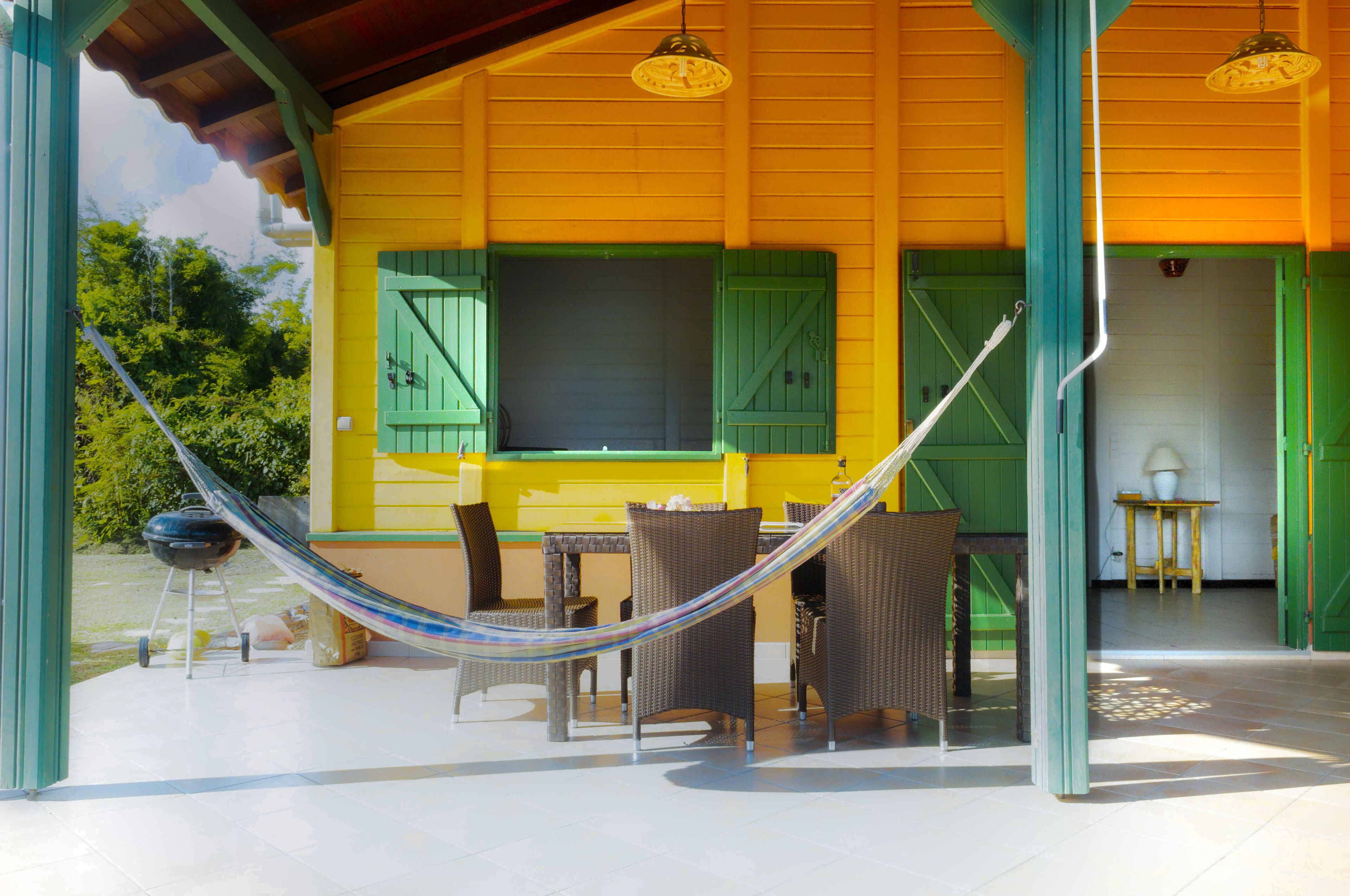 A covered outdoor area with a dining table and chairs, a hammock, and a view of a building with green shutters.