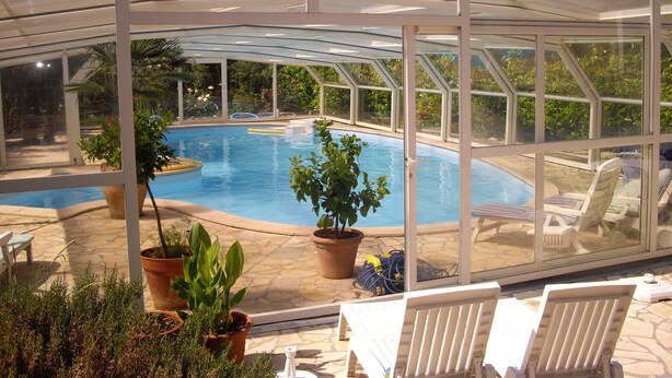 Ein überdachter Pool mit Glasverkleidung, umgeben von Blumentöpfen und Liegestühlen.