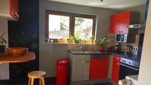 A modern kitchen with red and grey cabinets, a wooden stool, a red trash bin, and a window with potted plants. - A trip into nature (Bollendorf)