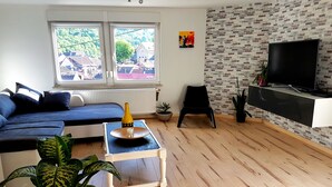 A modern living room with a blue sofa, a small table with a glass, and a TV on a brick-patterned wall. - A trip into nature (Bollendorf)