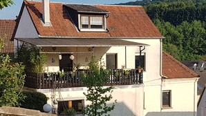 A two-story house with a balcony, a small garden, and a view of a forested hill. - A trip into nature (Bollendorf)