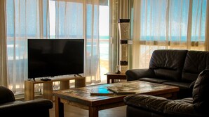 A living room with a large flat-screen TV on a wooden stand, a leather sofa, a coffee table with a book, and sheer curtains with a view of the outdoors. - Waterfront villa with swimming pool. Exceptional view (saline les bains)