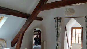 A bedroom with a large bed, a mirror, a window with curtains, and a wooden ceiling beam.
