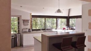 A modern kitchen with a central island, bar stools, and large windows offering a view of greenery.