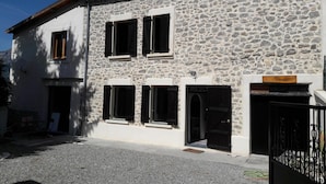 A two-story stone building with multiple windows and doors, a gravel driveway, and a metal gate. - EXPOSED STONE HOUSE (Ancelle)