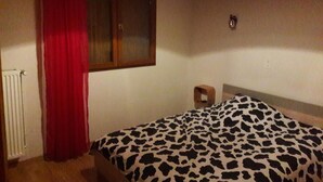 A bedroom with a bed, a window with curtains, a radiator, and a wall-mounted light fixture. - EXPOSED STONE HOUSE (Ancelle)