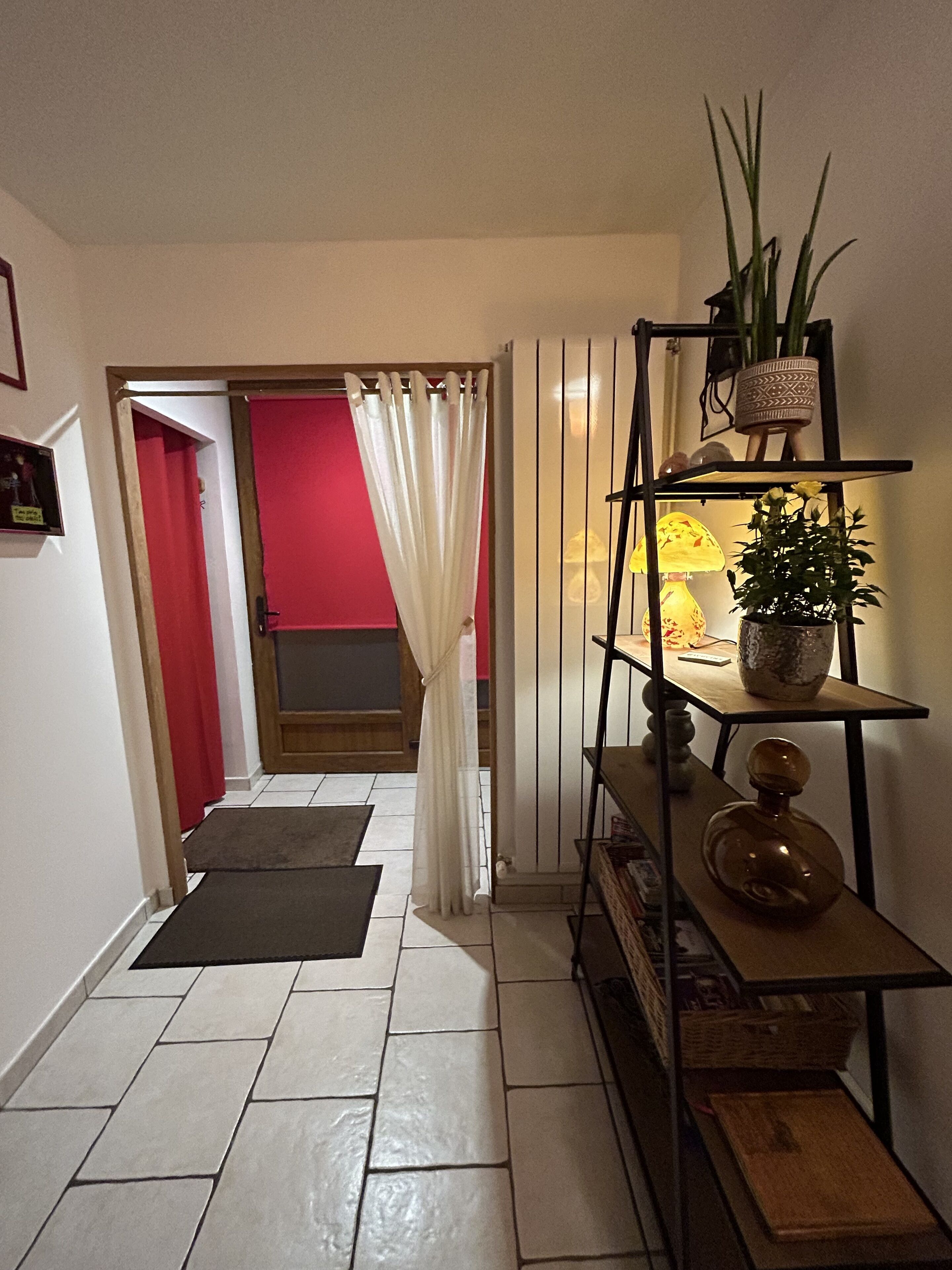 A hallway with a tiled floor, a white door with a red interior, a white curtain, a shelf with plants and a lamp, and a small table with a vase.
