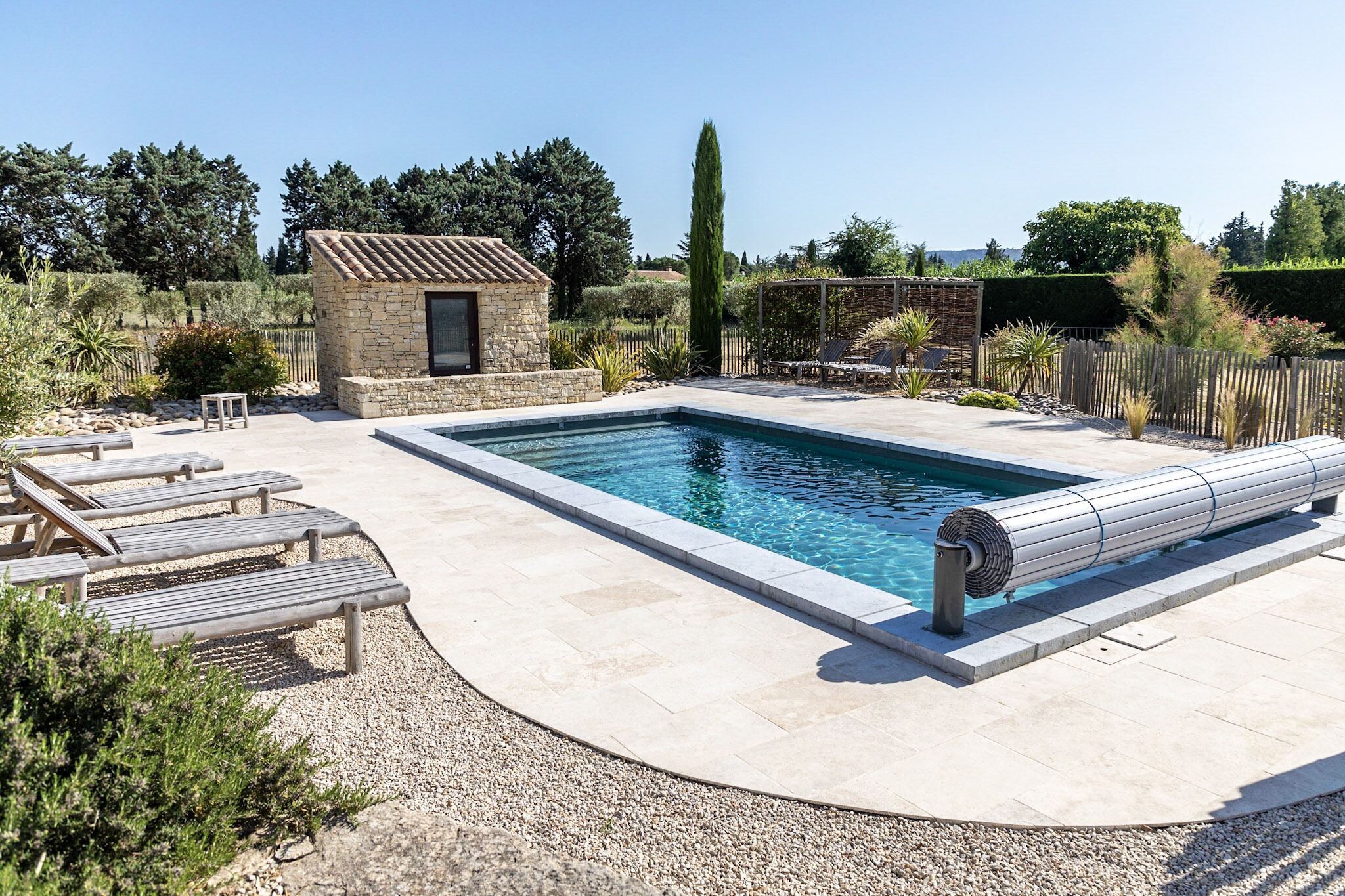 Une piscine rectangulaire bordée d’une terrasse en pierre, entourée de chaises longues et d’un petit bâtiment en pierre.