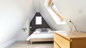 A bedroom with a sloped ceiling, a bed, a wooden dresser, a small table, and a window with a view. - Villa Joseph **, Apartment 4 people in Kaysersberg - 2 bedrooms (Kaysersberg-Vignoble)