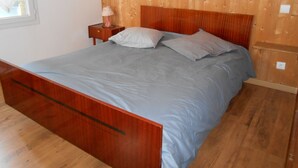 A bedroom with a wooden headboard, a bed with grey bedding, a nightstand with a lamp, and a window with a curtain. - 4-seater apartment 50 m2 (Laruns)