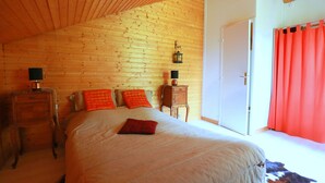 Un dormitorio con techo de madera, una cama con almohadas anaranjadas y rojas, una mesita de noche con una lámpara y una puerta con cortina roja.