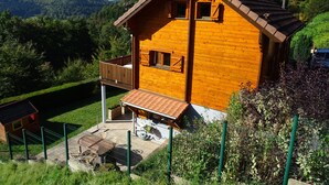 A wooden cabin with a balcony, surrounded by greenery and hills.