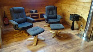 A cozy room with wooden walls, a skylight, two dark armchairs, a small ottoman, a radio, and a fire extinguisher.