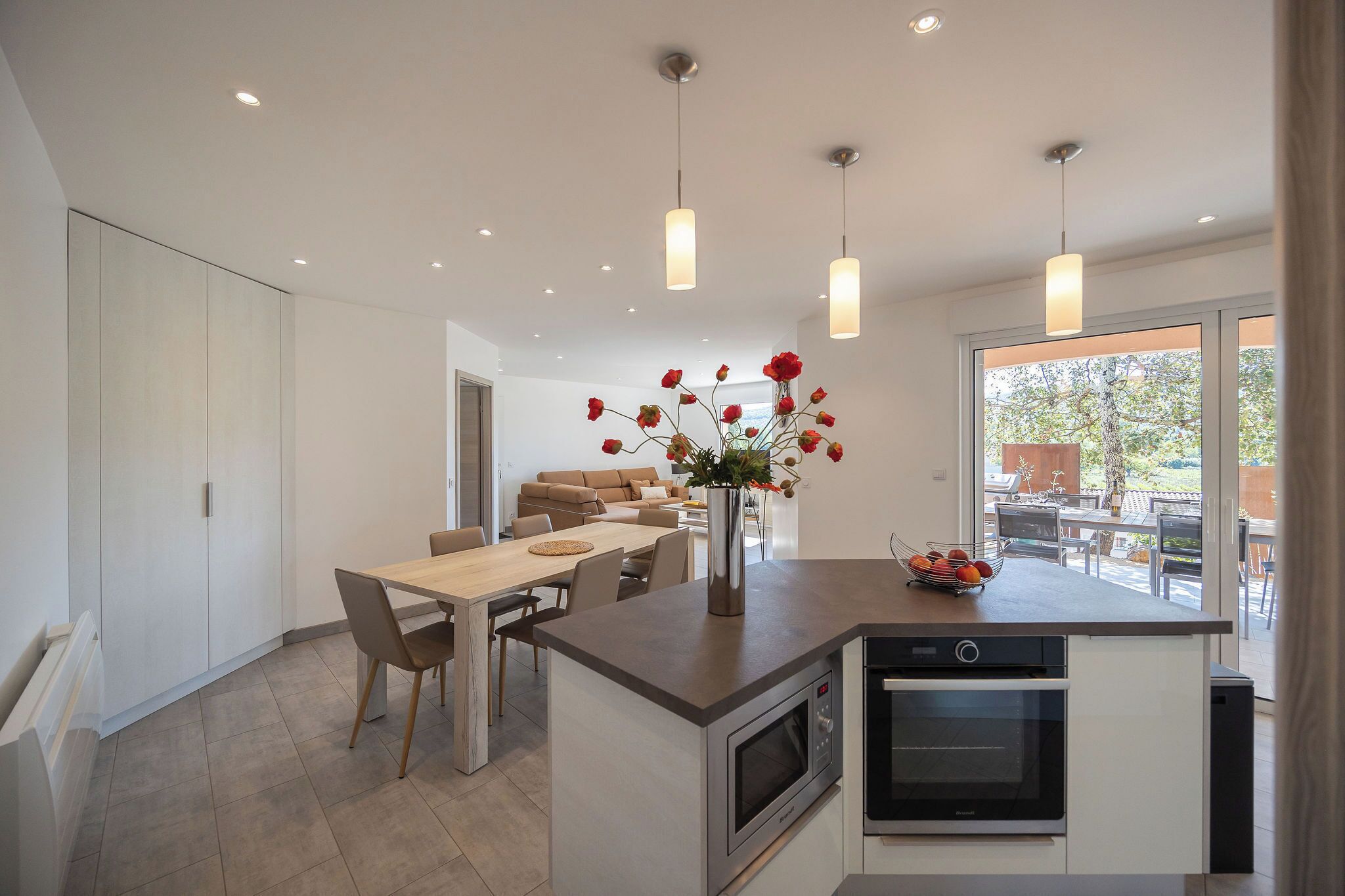 A modern kitchen with a central island, a dining area with a wooden table and chairs, and a large window providing a view of the outdoors.