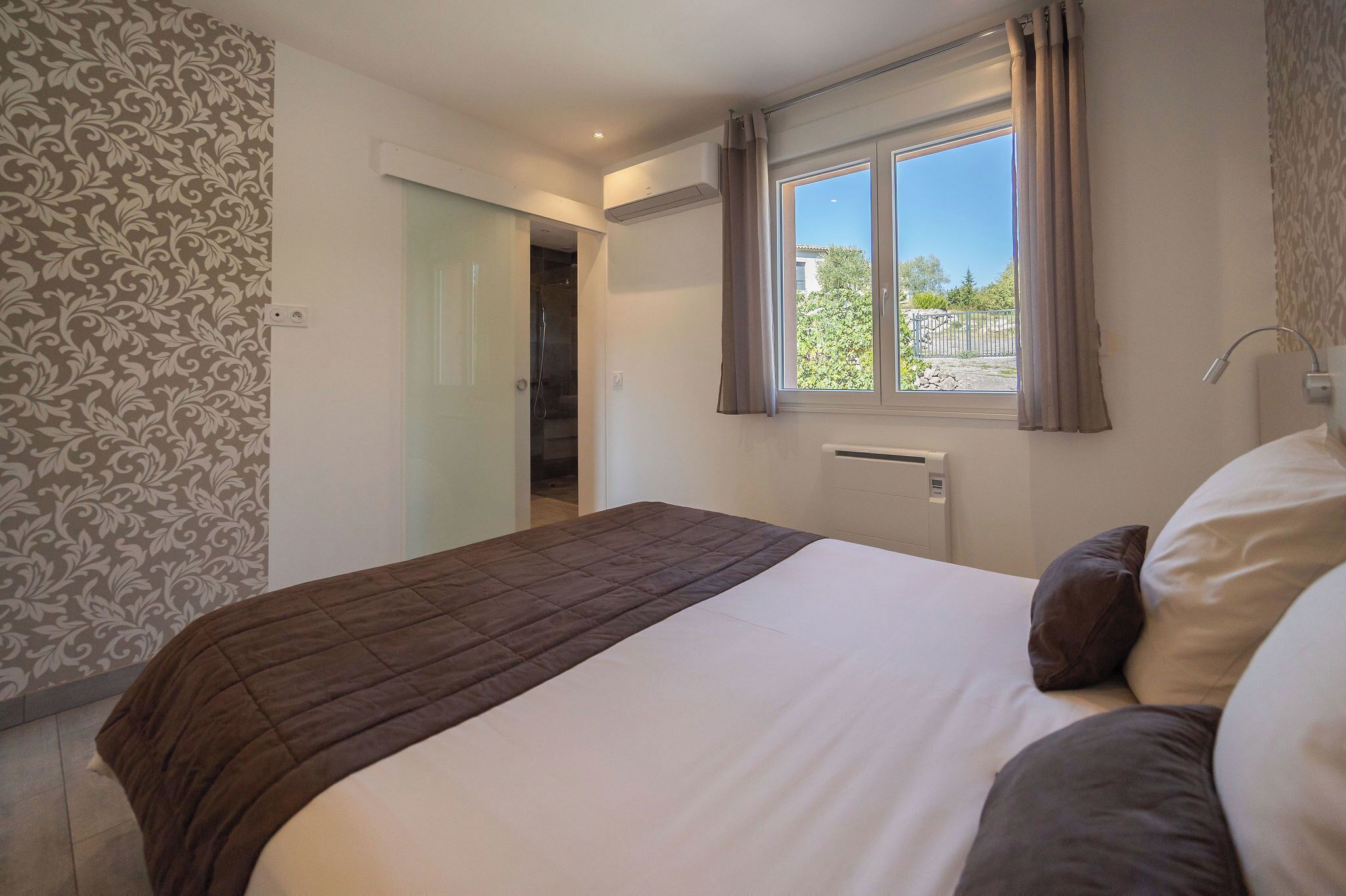 A bedroom with a bed, a window with curtains, a radiator, and a door leading to another room.