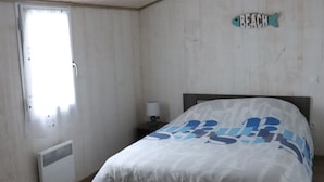 Un dormitorio con techo de madera, una cama con colcha blanca y azul estampada, una mesita de noche con lámpara y una ventana con vistas al exterior.