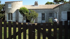 Un bâtiment rustique en pierre, avec un toit de tuiles, une clôture en bois au premier plan et une personne qui marche au loin.