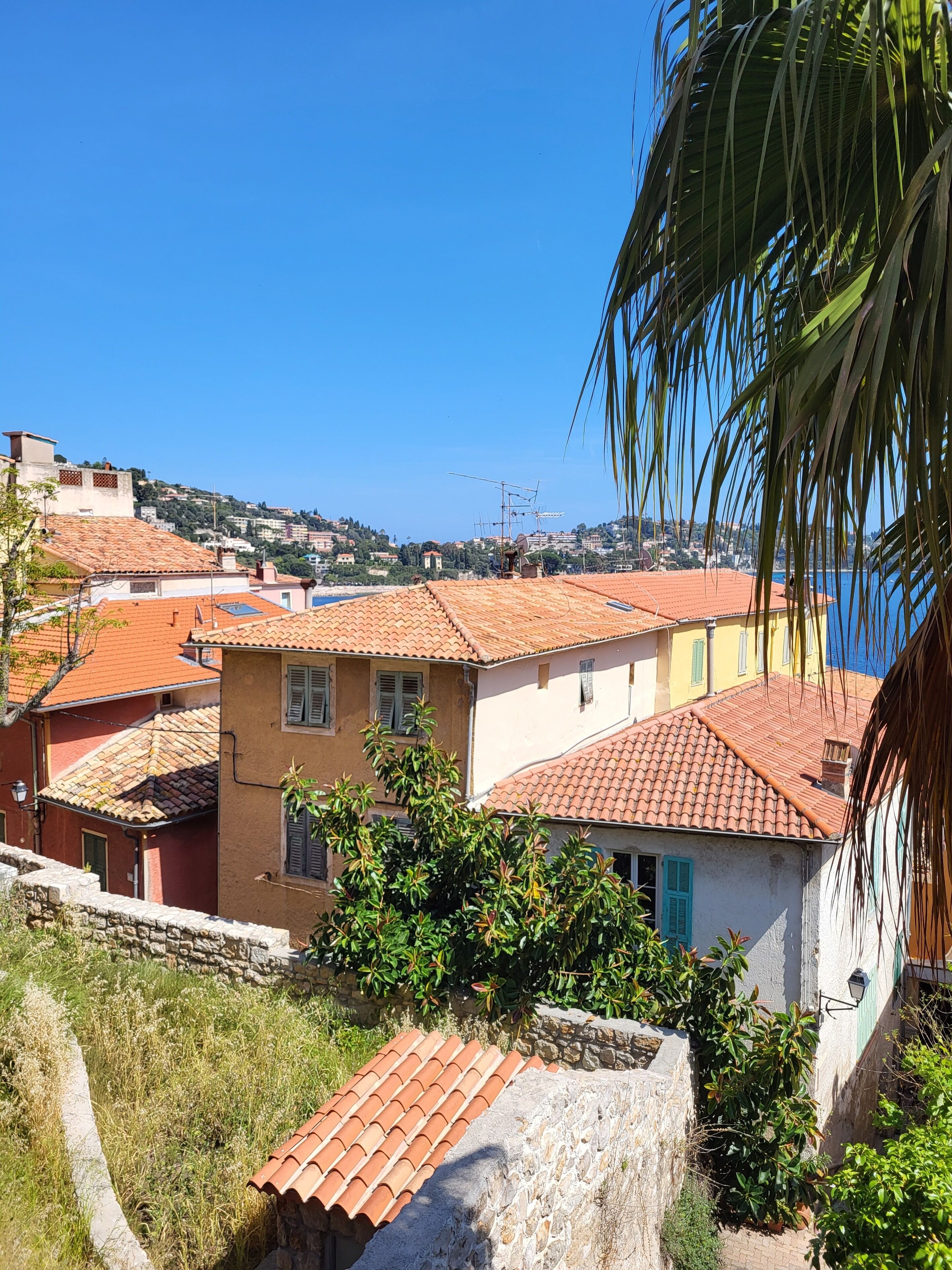 A coastal town with terracotta-tiled buildings, a stone wall, and a clear blue sky.