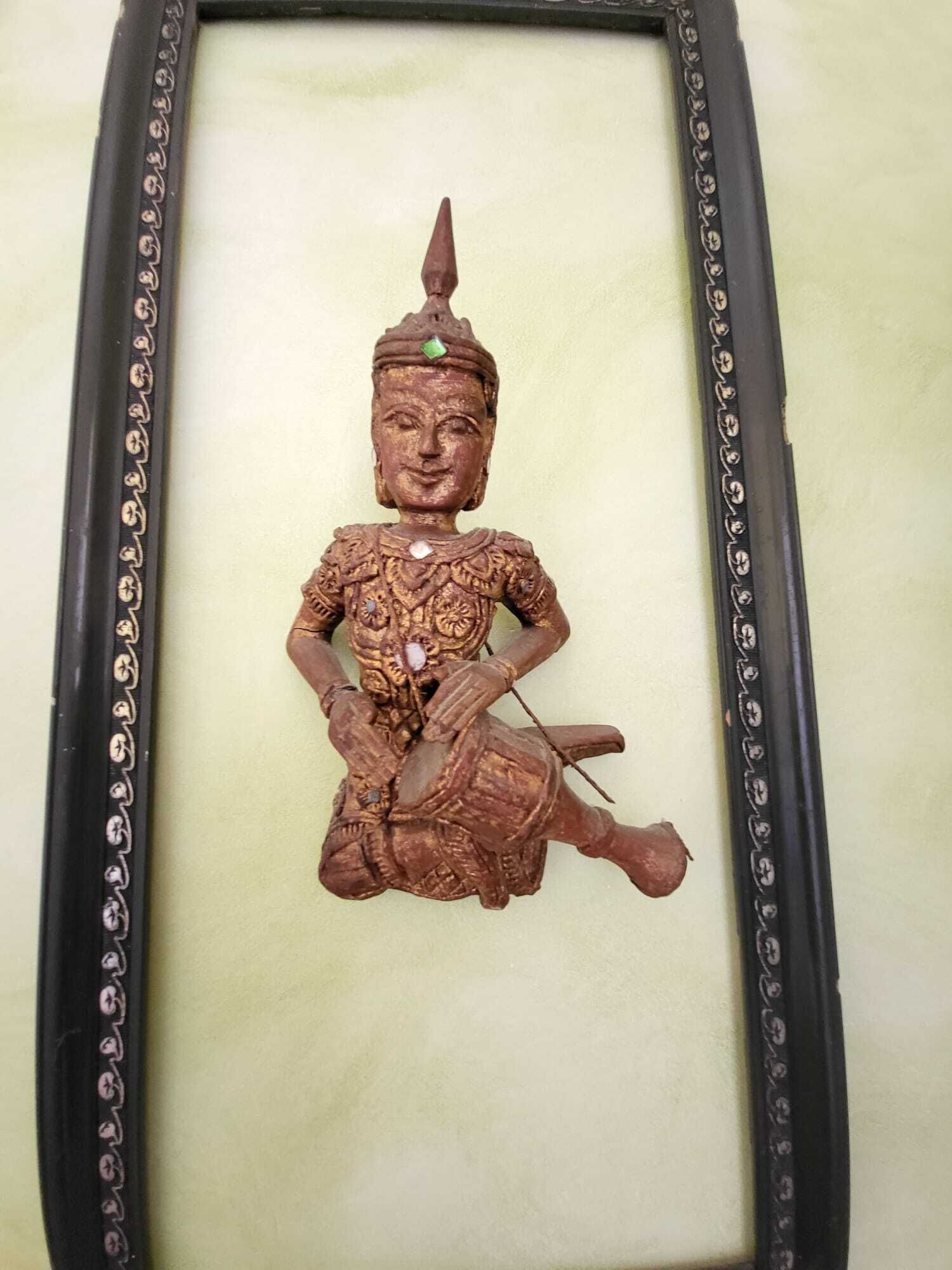 Une sculpture encadrée représentant une figure assise, ornée de motifs complexes, placée sur un fond uni.