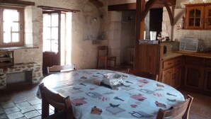 A rustic kitchen with stone walls, wooden beams, and a tiled floor. There is a round table with a patterned tablecloth, wooden chairs, and a window with curtains.