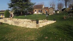 Un mur de soutènement en pierre, agrémenté d’un petit jardin, une maison en bois avec un toit rouge et un grand arbre en arrière-plan.
