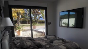 A bedroom with a large bed, a TV mounted on the wall, and a view of a garden through the sliding doors. - VILLA LES GRANGES- SWIMMING POOL surrounded by a green park overlooking the mountains  (Saint-Marcellin-Lès-Vaison)