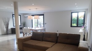 A modern living room with a sectional sofa, a dining area with chairs and a table, and large windows offering a view of greenery outside. - VILLA LES GRANGES- SWIMMING POOL surrounded by a green park overlooking the mountains  (Saint-Marcellin-Lès-Vaison)