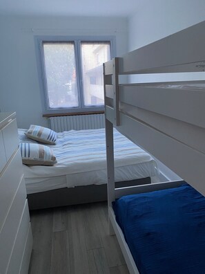 Une chambre avec un lit superposé, une fenêtre avec des stores, un radiateur et un plancher en bois.
