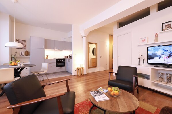 A modern living room with a wooden coffee table, black leather chairs, a television, and a kitchen in the background.