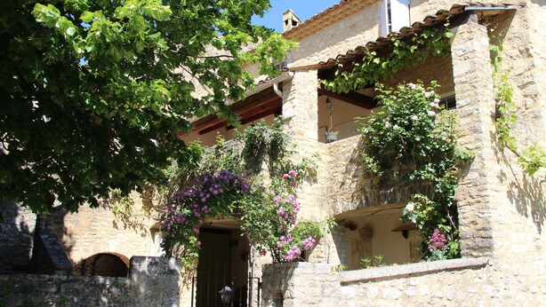 Un bâtiment en pierre doté d’une façade rustique, d’un balcon orné de plantes suspendues et d’une entrée bien visible.