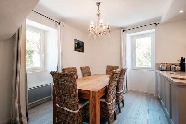 A dining area with a wooden table and wicker chairs, a chandelier, and windows with curtains. - La Maison du Tilleul _ charming house with heated pool from April to October (La Roque-Sur-Cèze)