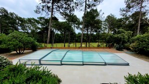 En swimmingpool med glasvægge og en træterrasse hele vejen rundt, beliggende i en anlagt have med forskellige buske og træer.