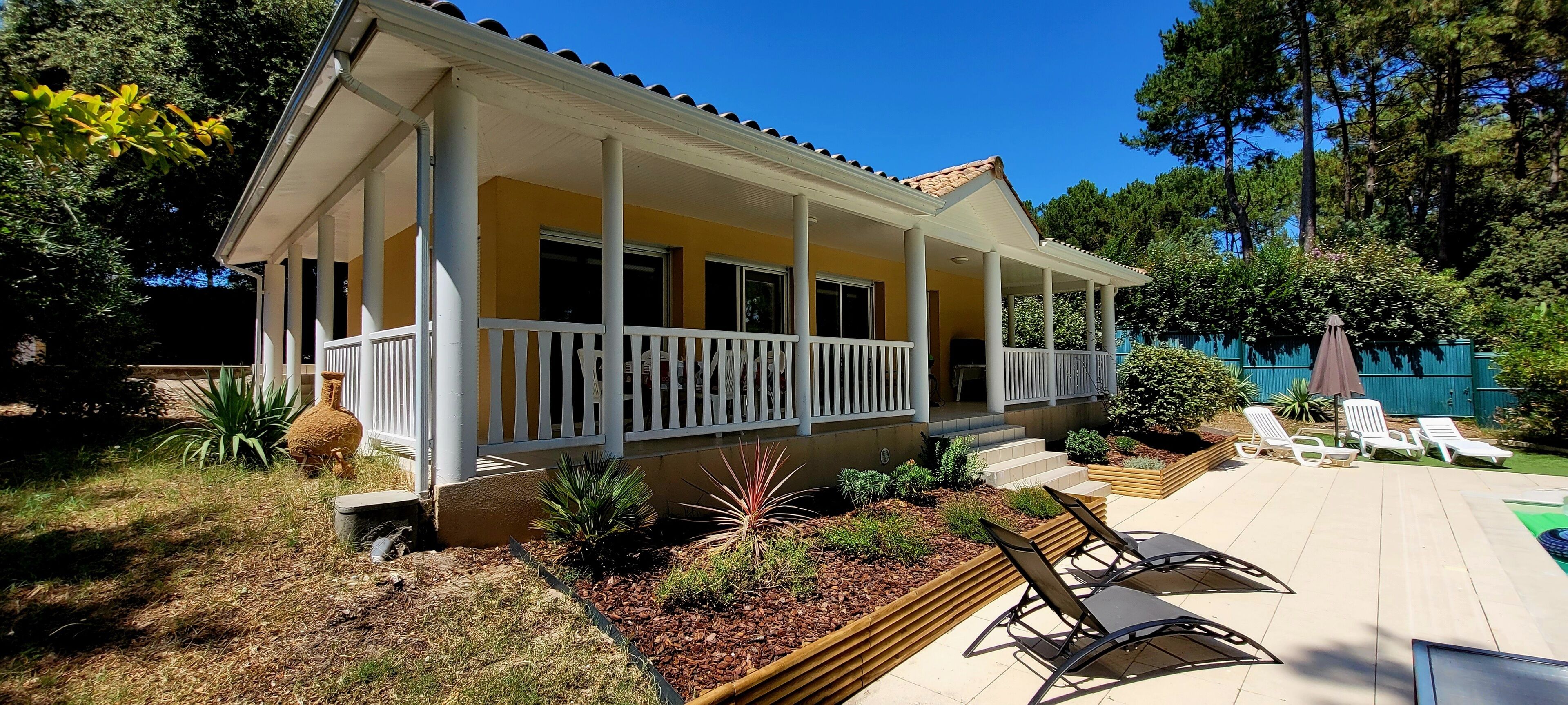 Ein zweigeschossiges Haus mit überdachter Veranda, einer Terrasse mit Liegestühlen und einem Garten mit verschiedenen Pflanzen.