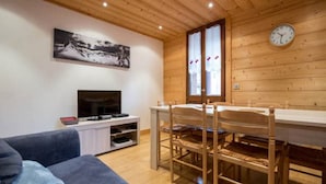 L’intérieur chaleureux d’un chalet avec un plafond en bois, une table à manger entourée de chaises, un téléviseur posé sur un meuble blanc et un tableau accroché au mur.