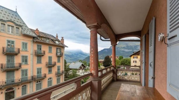 Un balcon avec une table en bois, d’où l’on peut admirer des bâtiments et des montagnes en arrière-plan.