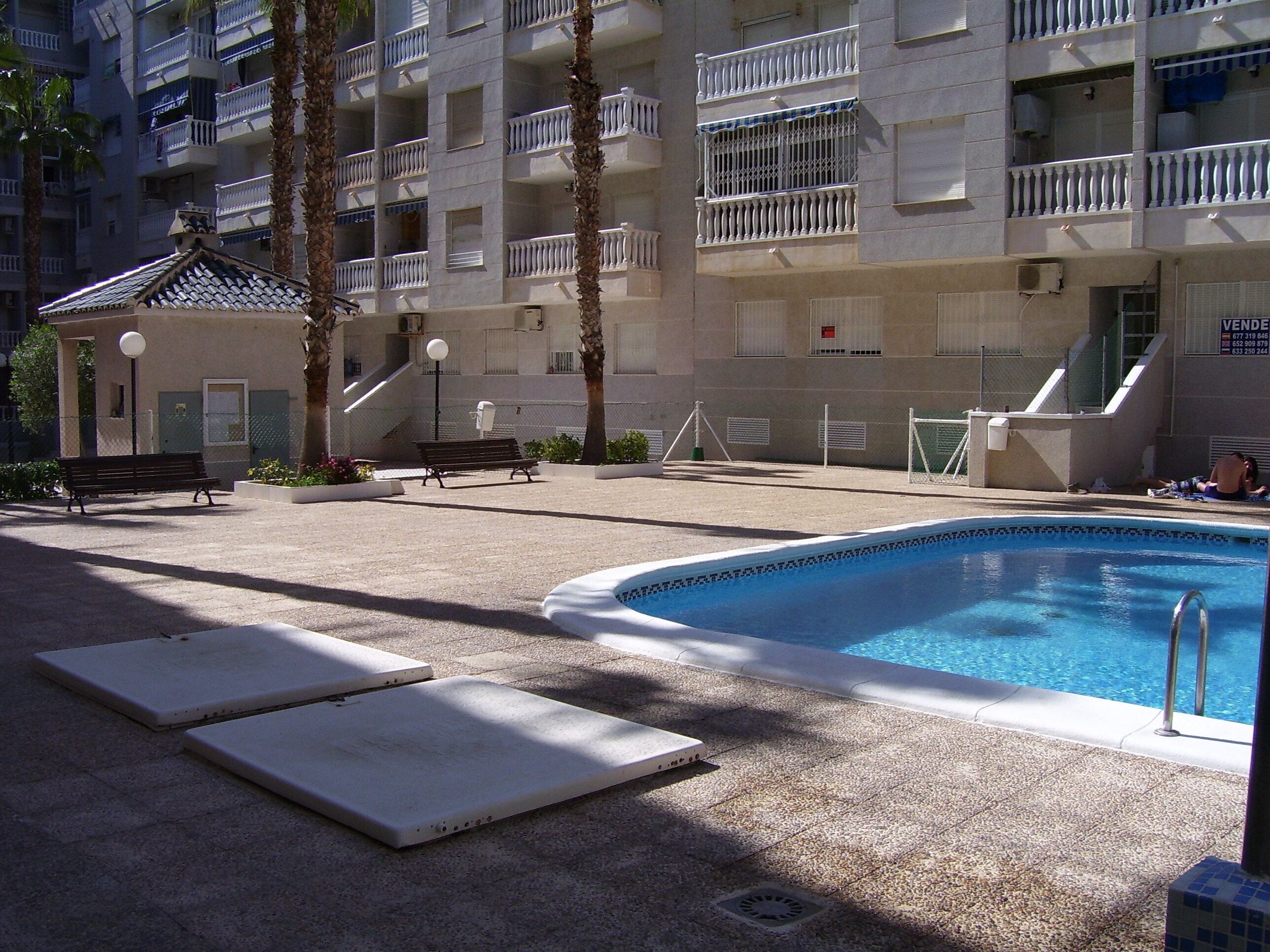 A pool area with lounge chairs and a bench, surrounded by a building with balconies and a covered seating area.